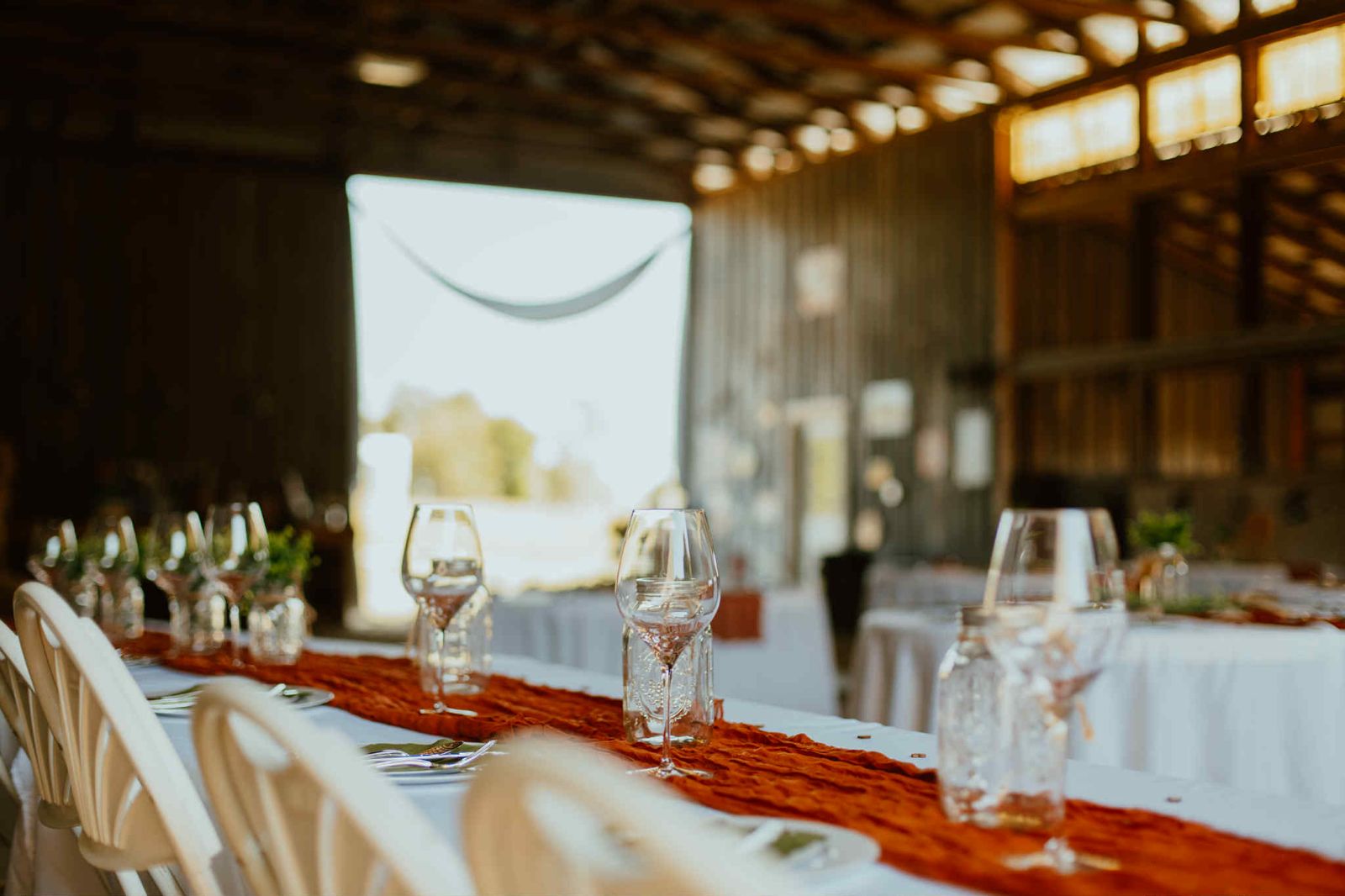 Long reception table set with wine glasses and a rust table runner, barn doors open to the evening light