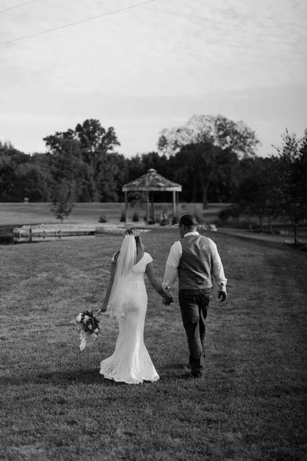 Bride and groom walking hand in hand across the lawn toward the pond gazebo, black and white